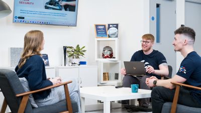 Sam, James and Richard gathered round a coffee table, focused on a laptop, collaborating together.