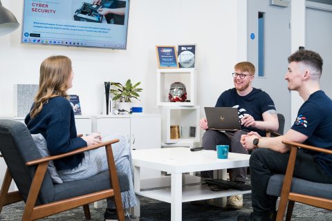 Sam, James and Richard gathered round a coffee table, focused on a laptop, collaborating together.