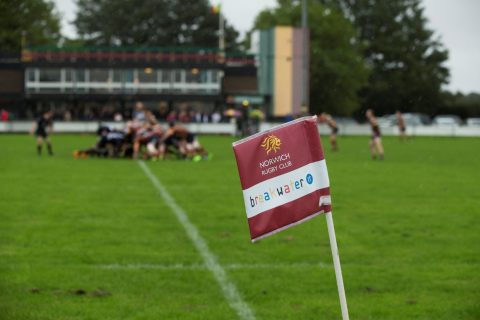 Norwich Rugby Club flag with Breakwater IT logo and rugby players in the background.