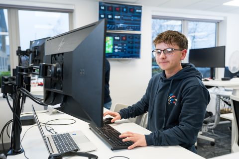 Alec is standing at a desk, working on his computer, with a keyboard ready for typing.