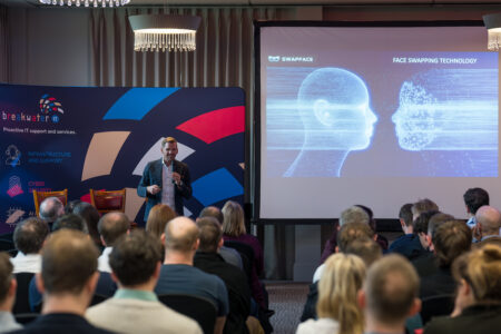 Conference room with rows of attendees sat listening to a speaker stood at the front. The speaker is stood in front of a large decorative banner. Next to the banner is a presentation screen with a slide depicting deep faking.