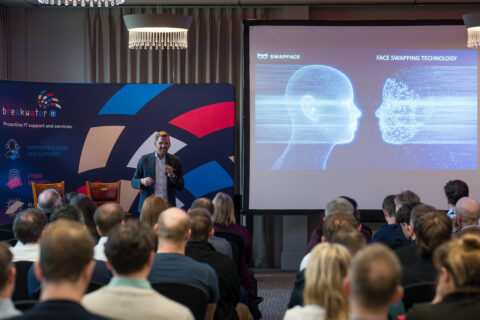 Conference room with rows of attendees sat listening to a speaker stood at the front. The speaker is stood in front of a large decorative banner. Next to the banner is a presentation screen with a slide depicting deep faking.