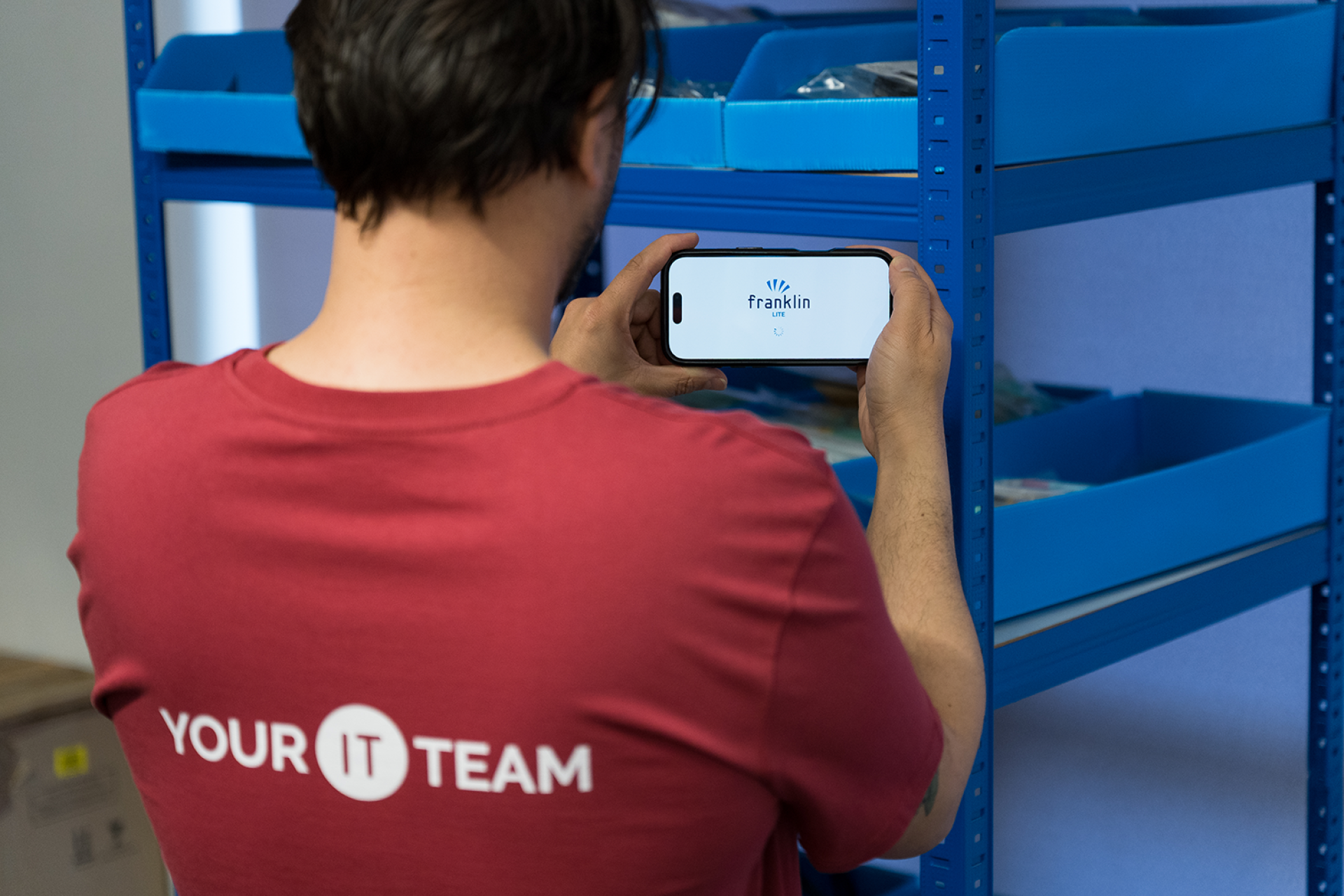 Man stood in stock room wearing red t-shirt that reads 'Your IT Team' across the back. He is holding a phone showing the Franklin logo.