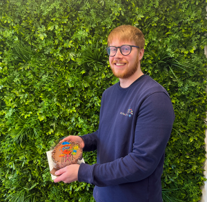 James Steers  holding a decorated cake in front of a greenery wall.